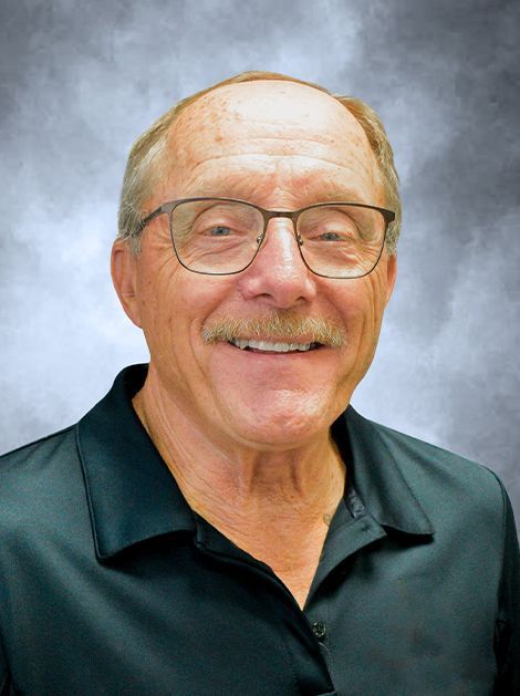 Smiling man wearing glasses and a black polo shirt against a blue-gray studio backdrop