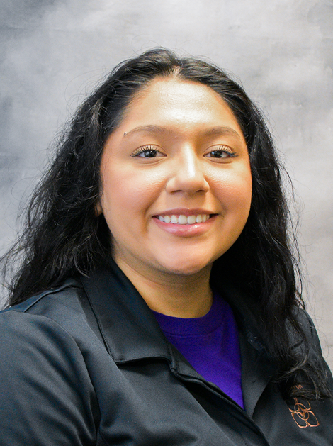 Smiling person in a dark jacket with long black hair against a gray studio backdrop