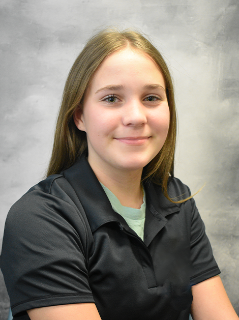 Person in a black polo shirt seated against a gray studio backdrop, smiling gently.