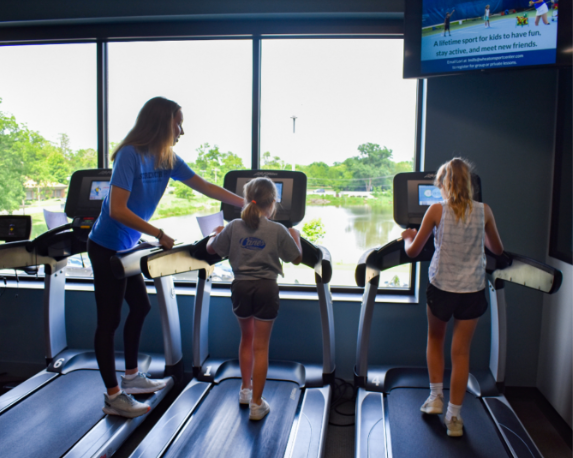 Three people using treadmills in a gym, with one instructor helping a child.
