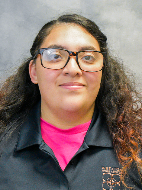 Portrait of a person wearing glasses, a black jacket, and a bright pink shirt against a gray backdrop.