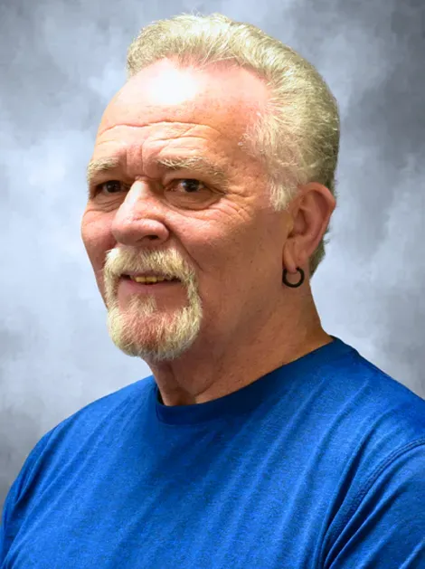 Portrait of a man with short blond hair and goatee, wearing a blue shirt against a gray backdrop