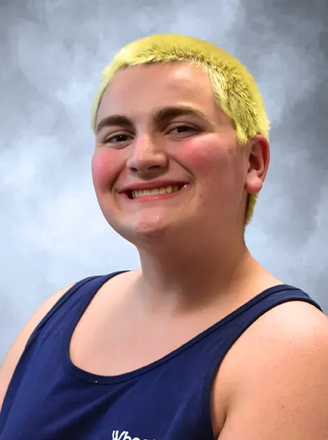 Smiling person with short blond hair in a navy tank top against a cloudy studio backdrop