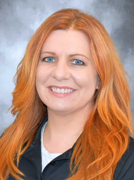 Smiling woman with long orange hair wearing a dark jacket against a gray studio background