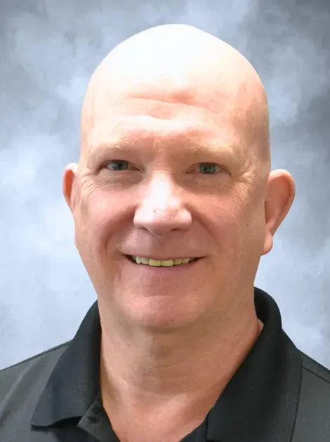 Smiling bald man in a black polo shirt against a blue-gray studio backdrop
