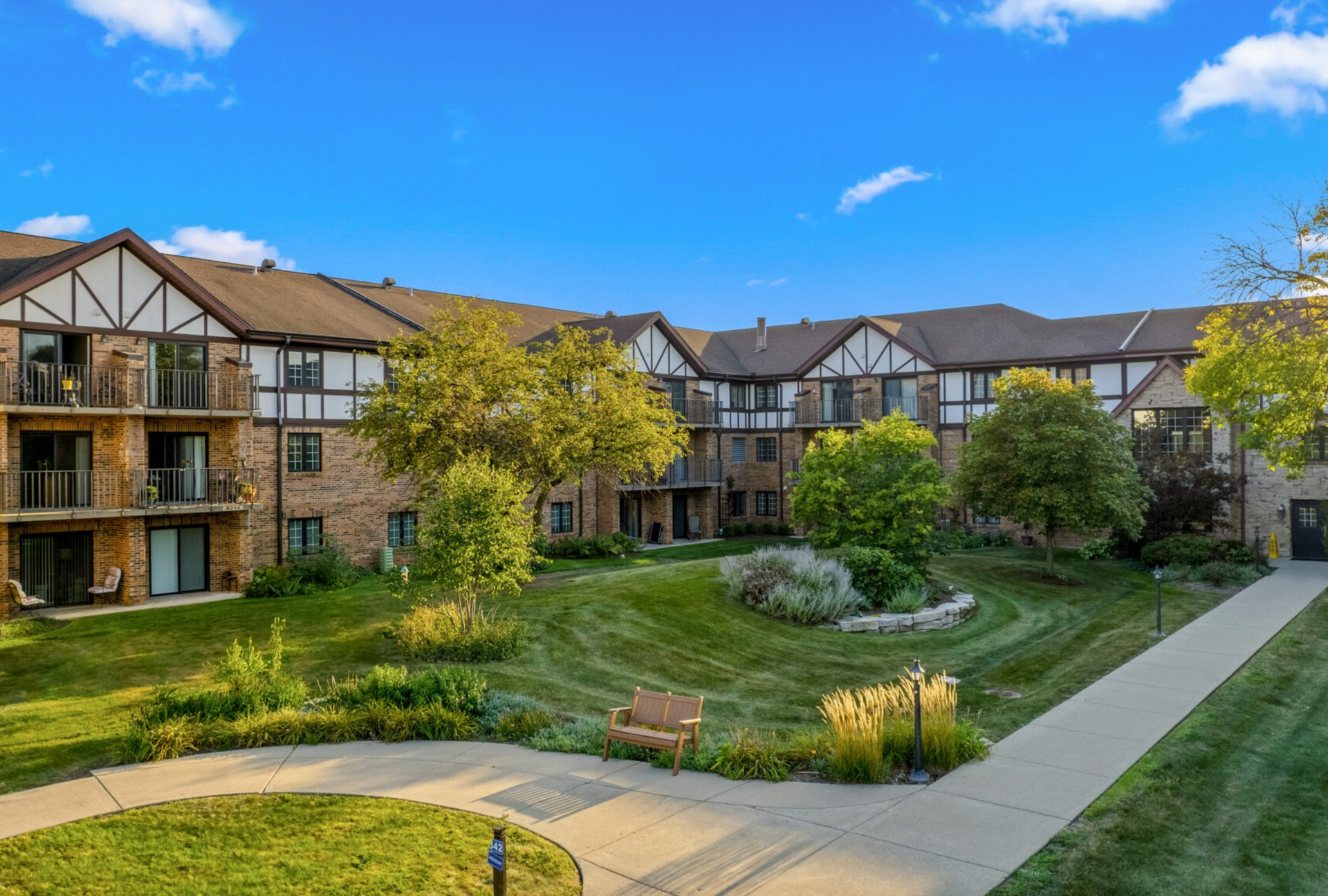 Courtyard of modern apartment buildings with green lawn, trees, and a bench under a blue sky