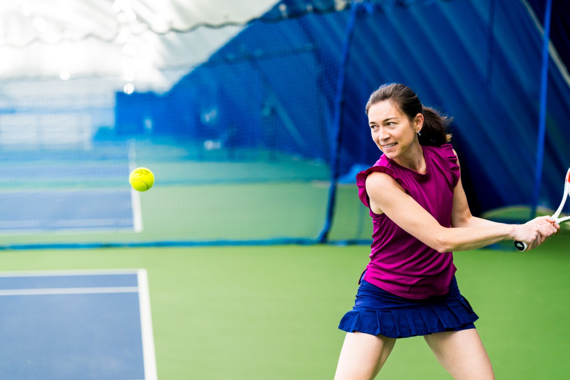 Tennis player in magenta shirt and navy skirt swings a racket at an outdoor court ball