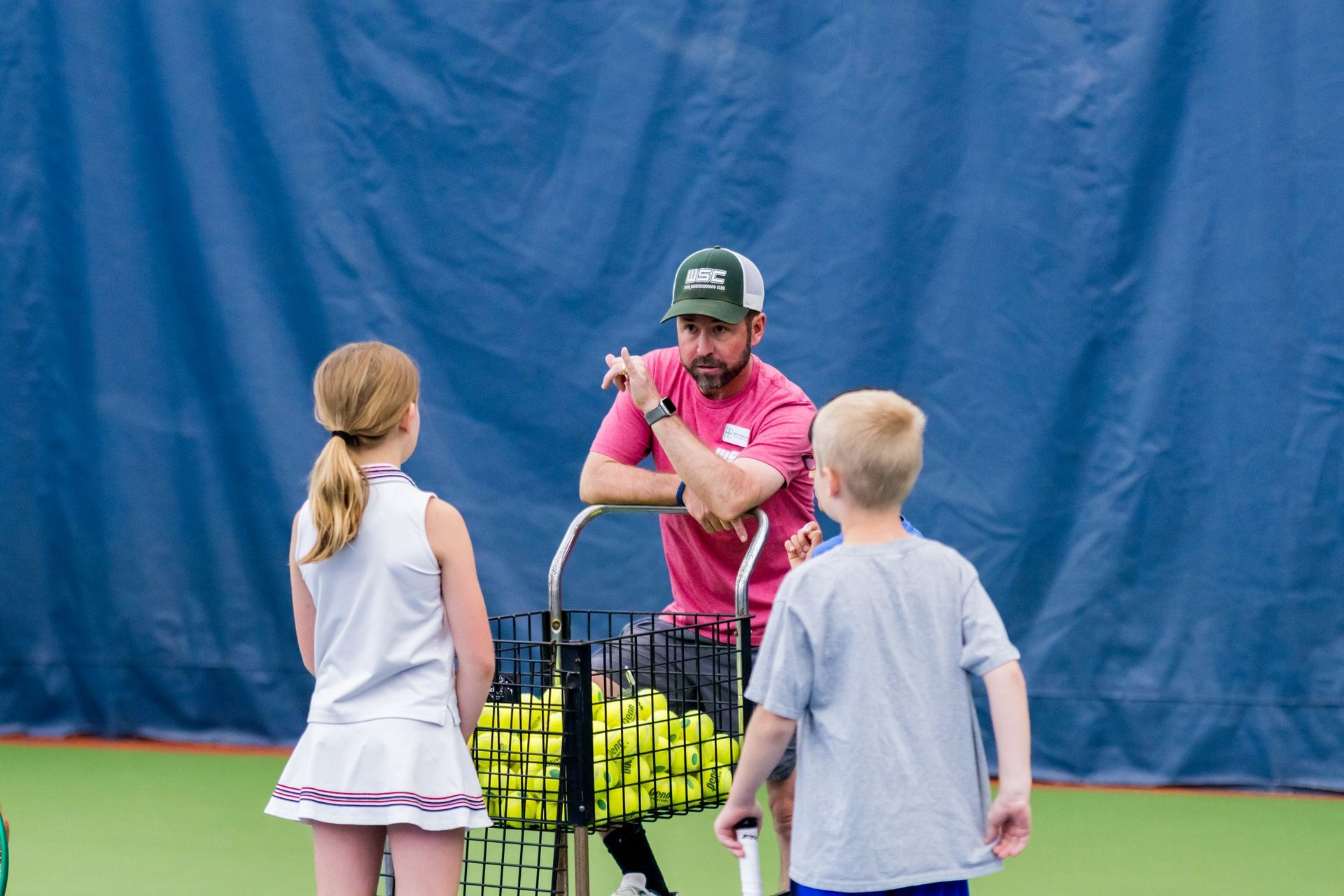 Coach in pink shirt showing two children a tennis drill beside a basket of balls outdoors