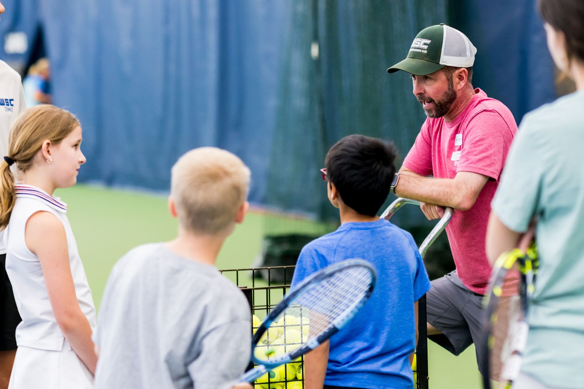 Coach talking to young tennis players on a court, with rackets and a blue fence in the background