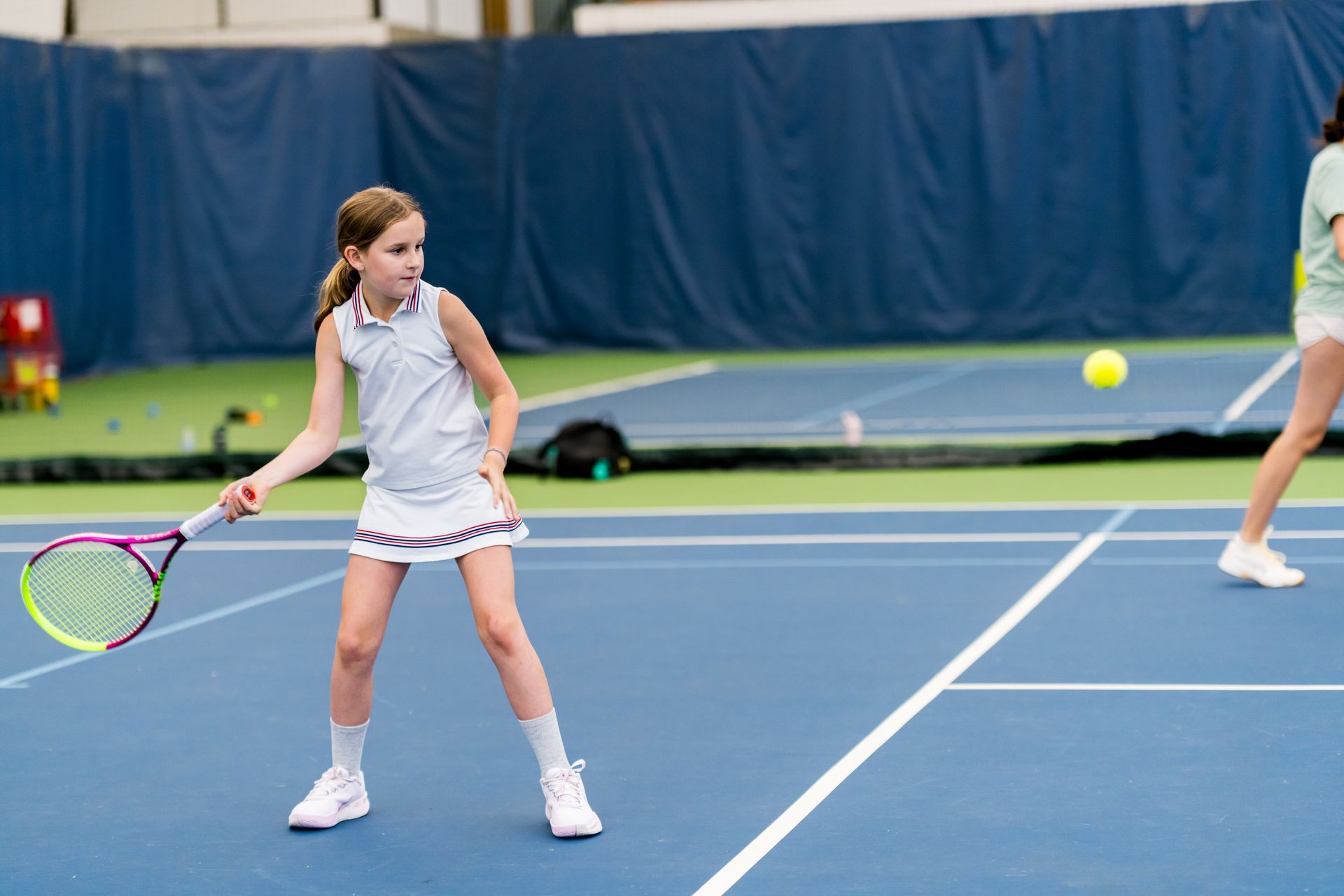Young tennis player in white swings a racket on an outdoor blue court with another player in the background