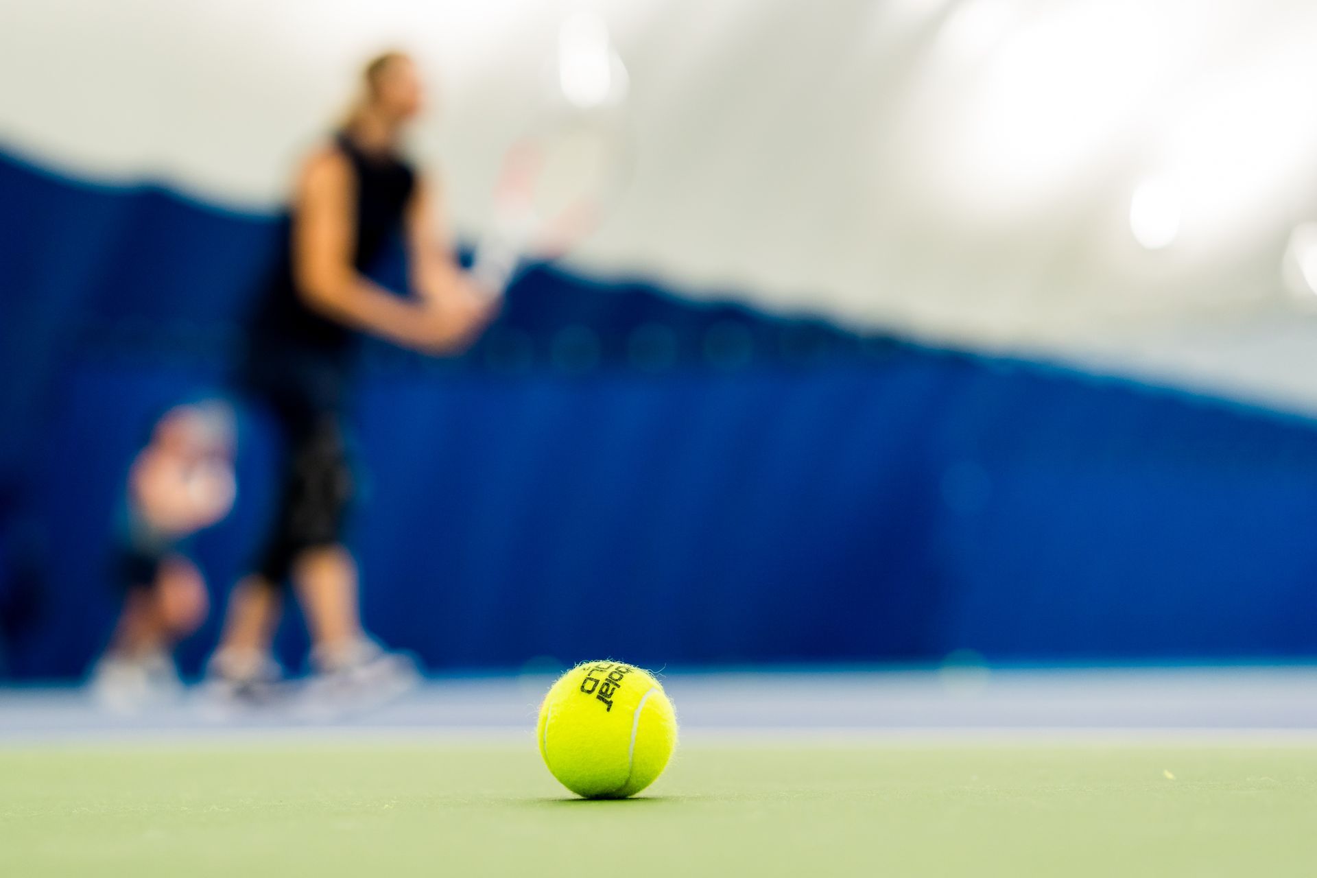 Tennis ball on court in foreground, with blurred player and child in the background