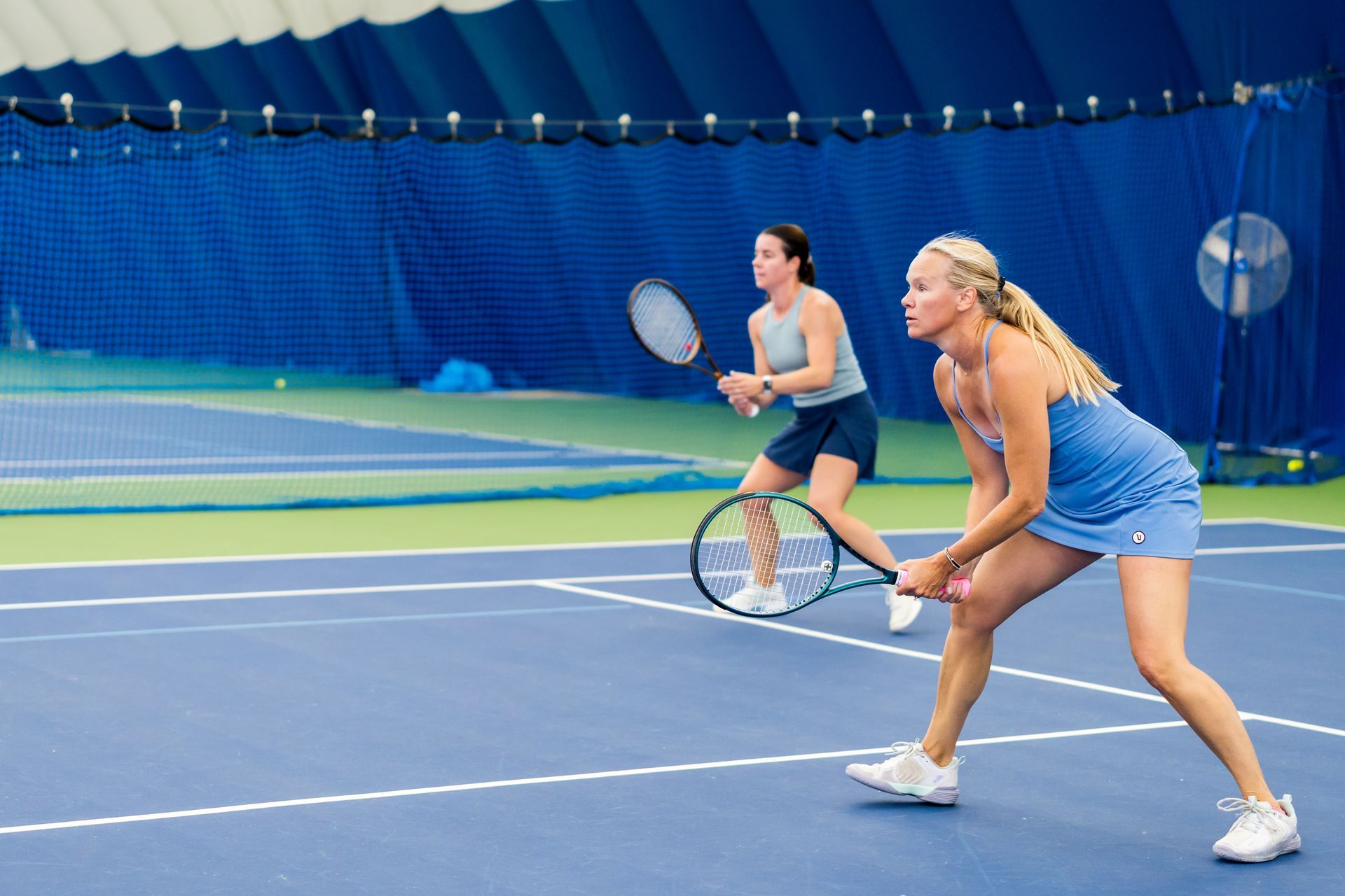 Two tennis players on an indoor court, one in blue ready to return a shot