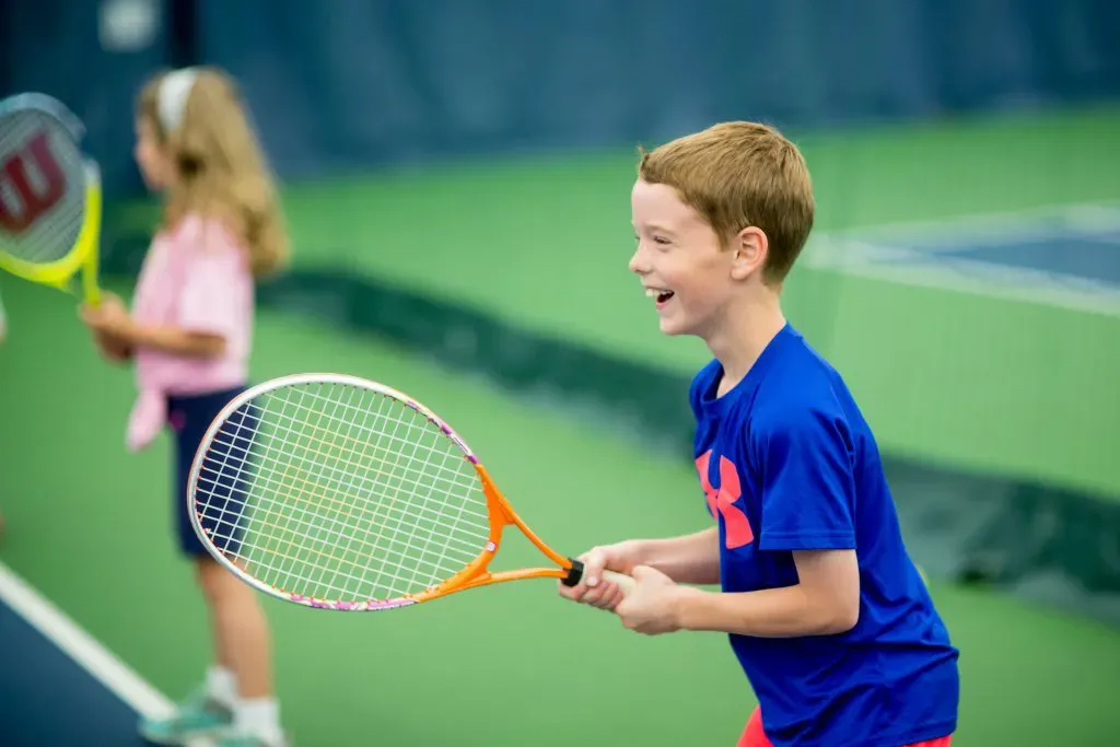 Boy in blue shirt playing tennis on an outdoor court, with another player blurred in the background