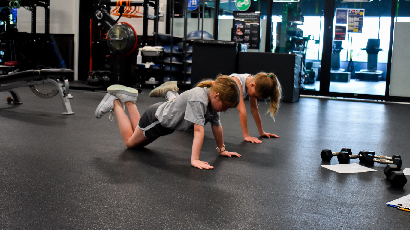 Two people doing push-ups on a gym floor with dumbbells nearby