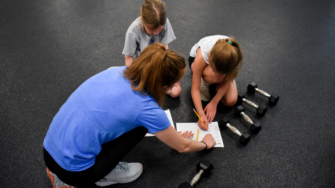 Three people kneel on a gym floor reviewing a paper near dumbbells.