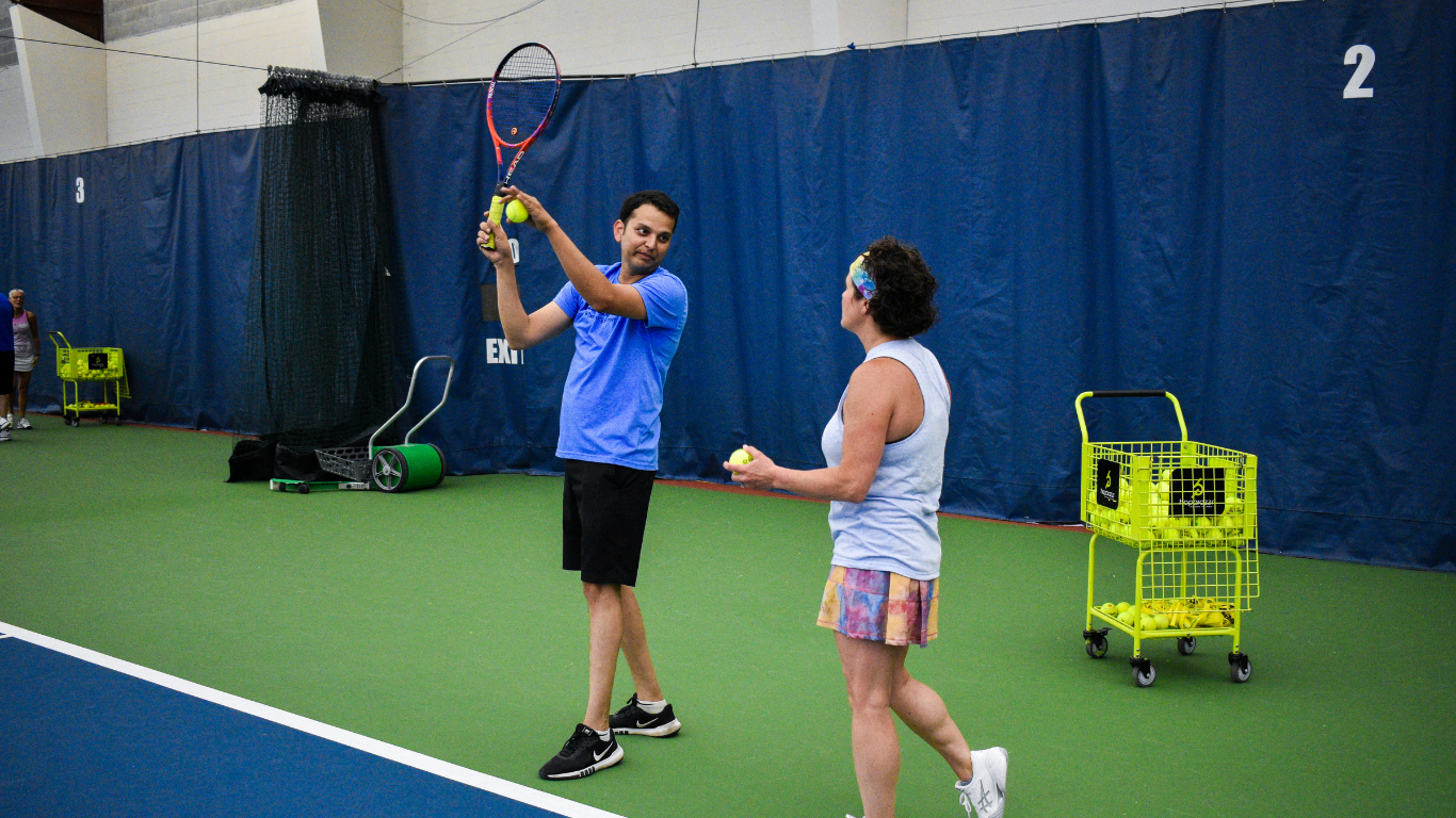 Two people on an indoor tennis court, one holding a racket near a basket of balls.