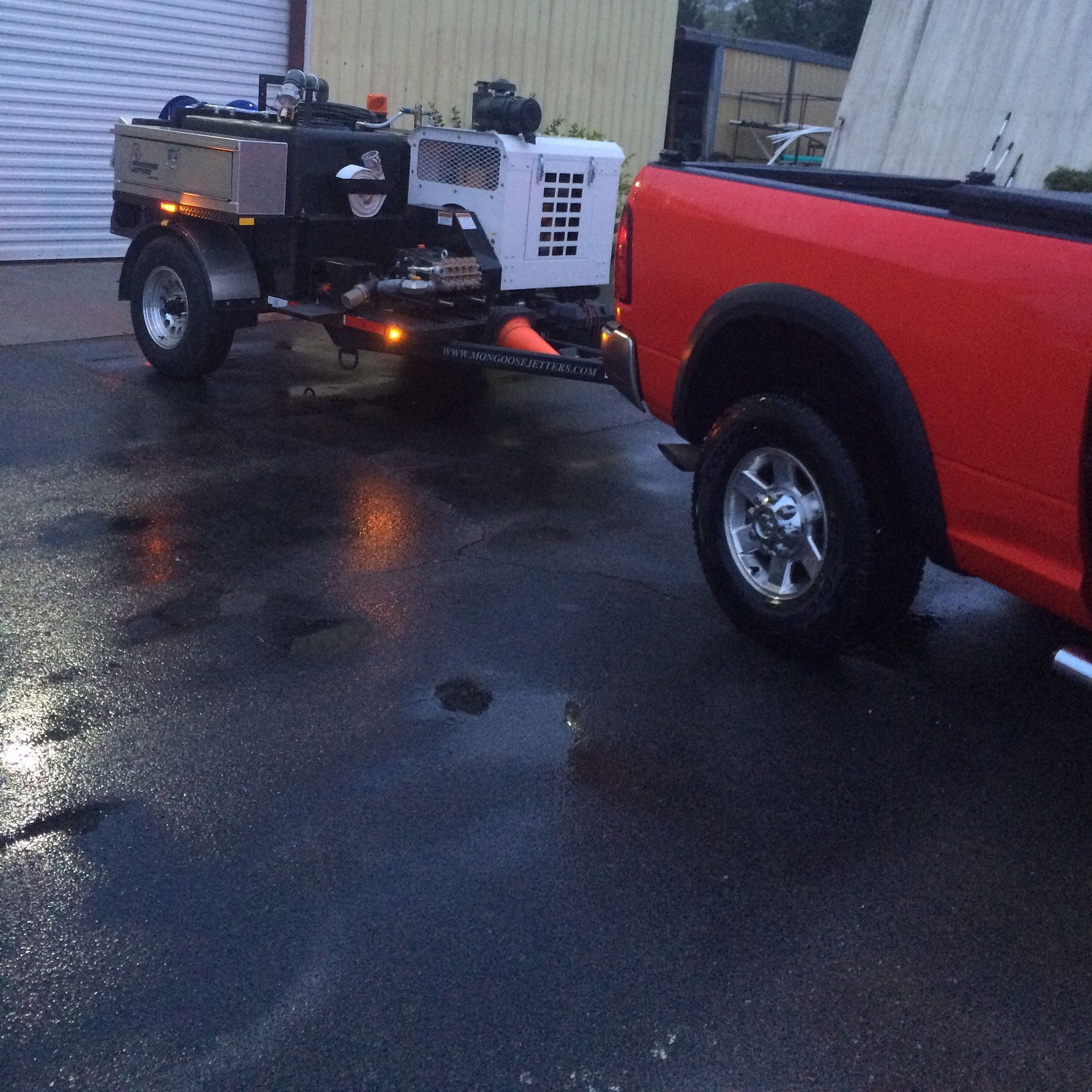 Red truck towing a trailer with road maintenance equipment on wet asphalt.
