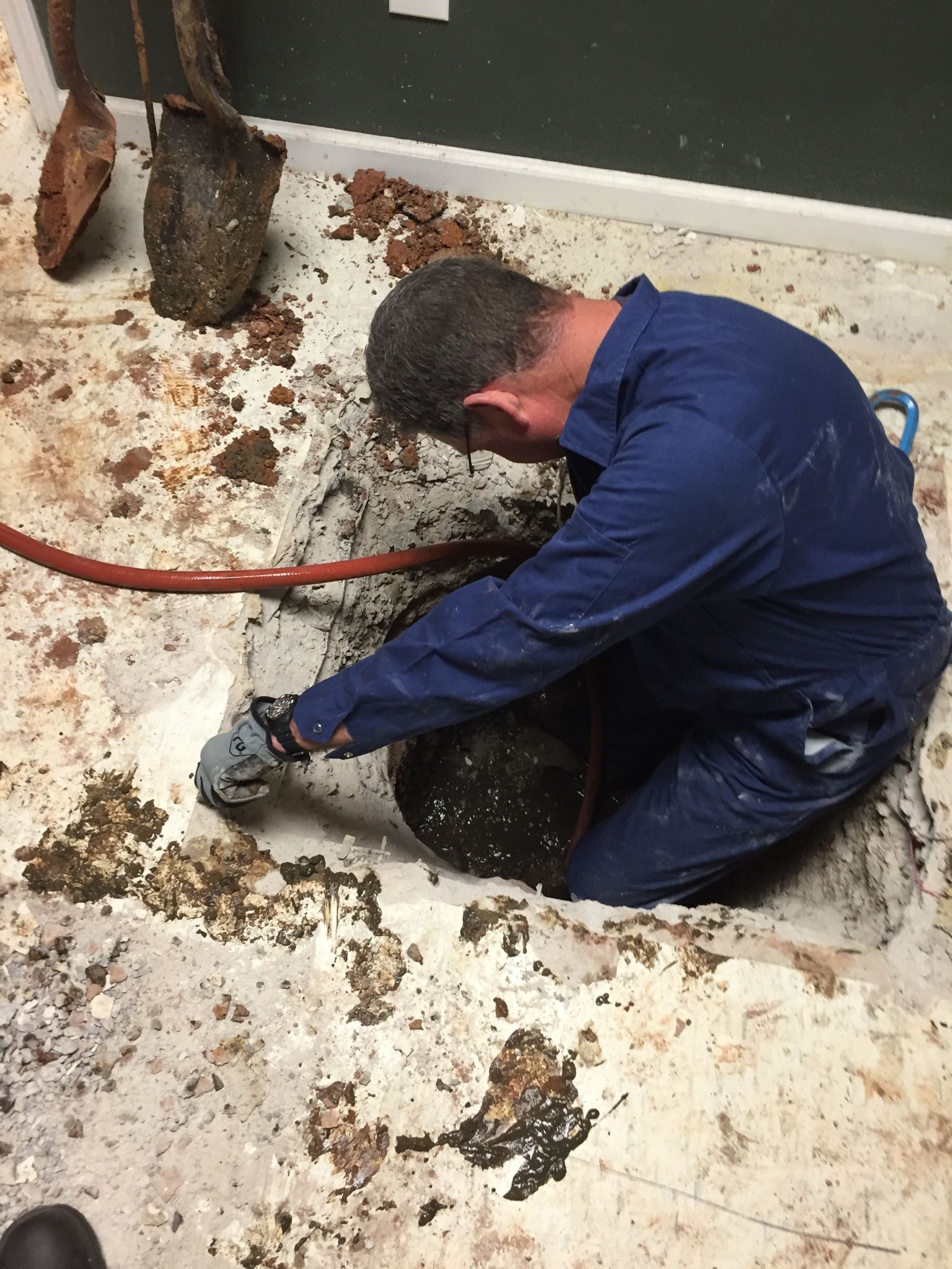 Man in blue jumpsuit working in an open hole in a dirty floor. He is handling something in the hole with a tool.