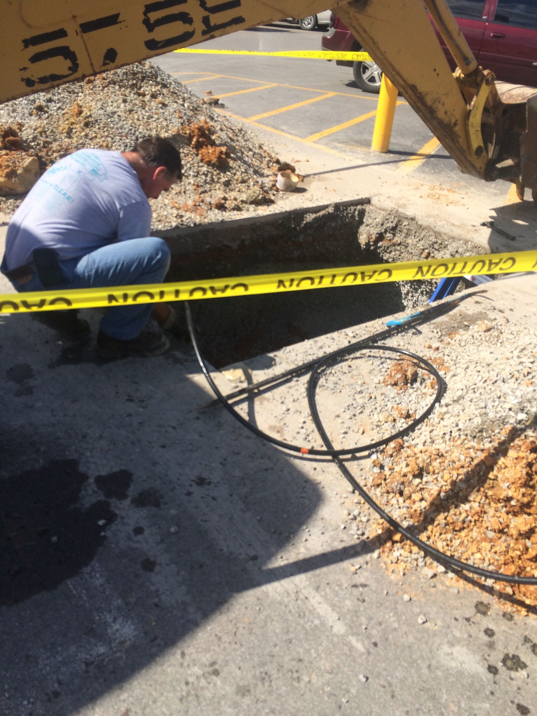 Man working at a hole in pavement, next to an excavator. Yellow caution tape around the site.