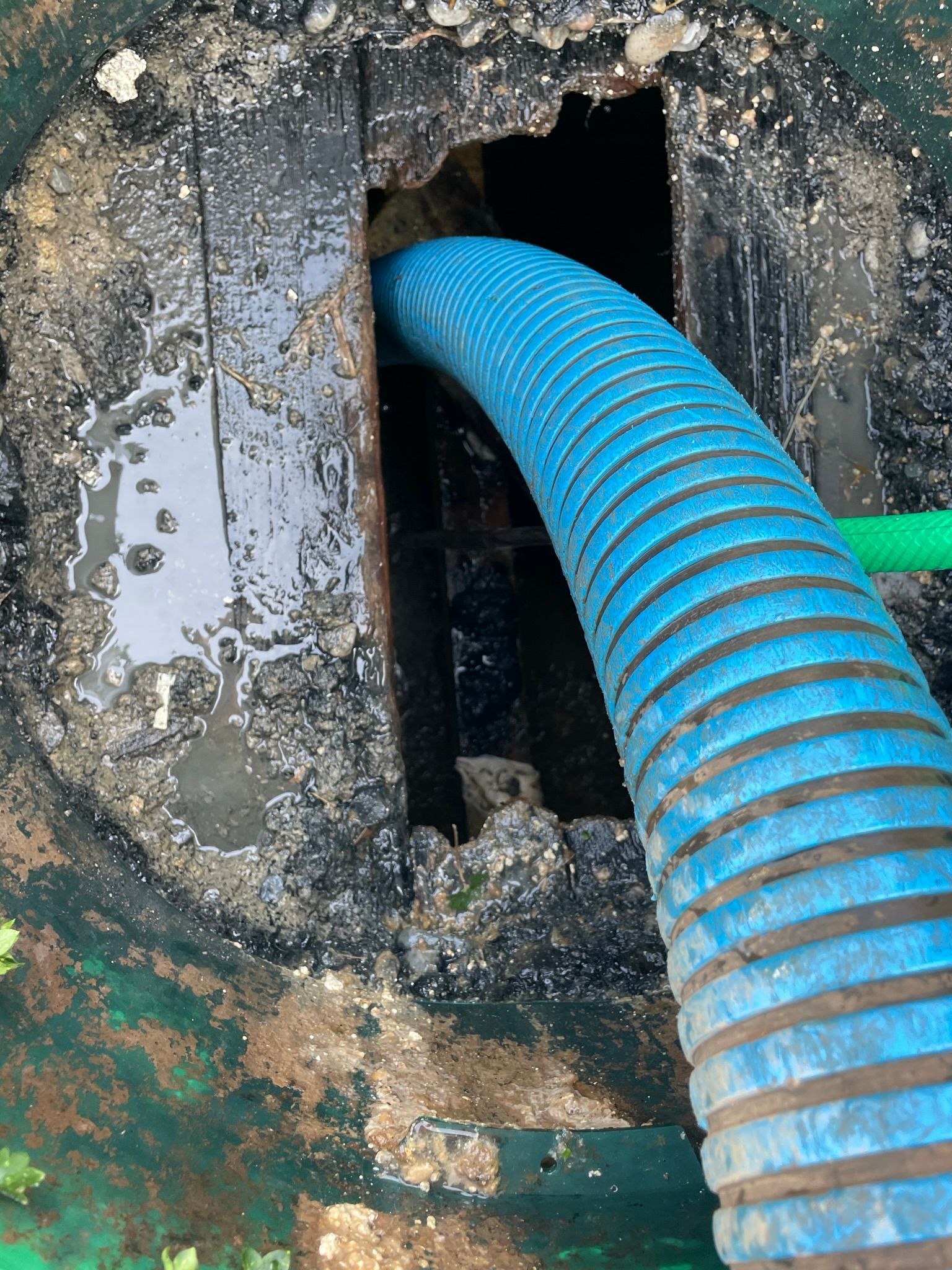 Blue hose inserted into a dark, open septic tank. Black sludge surrounds the opening.