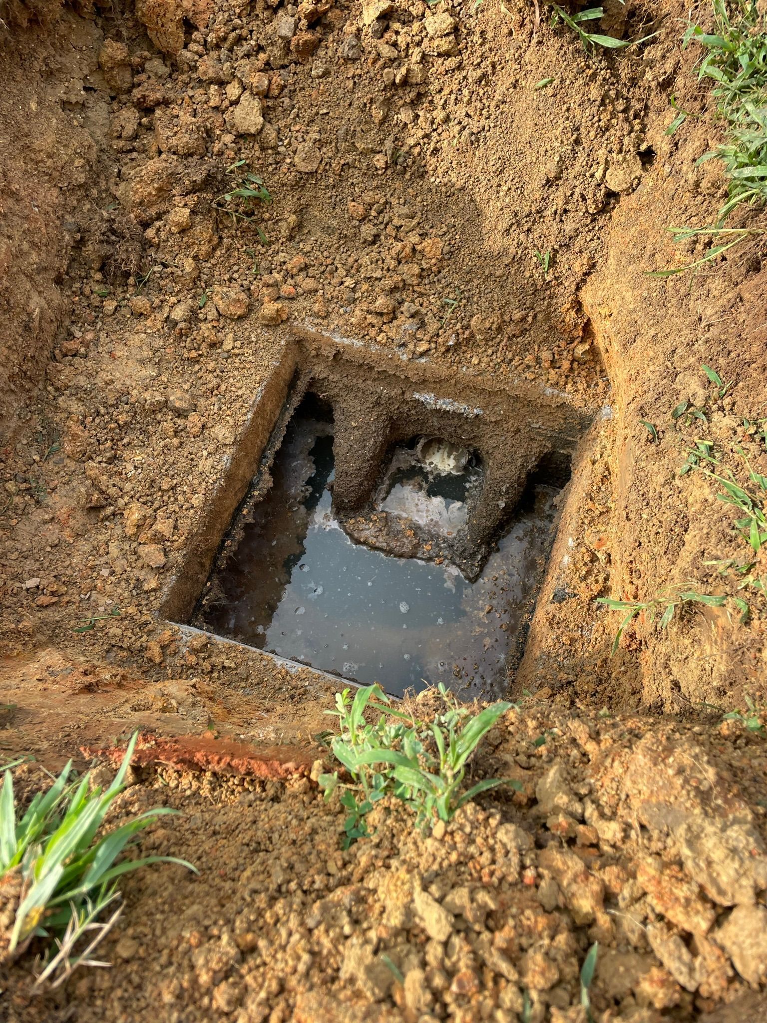 Square hole in dirt filled with murky water; exposed utility access point.