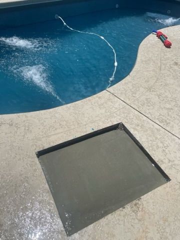 Concrete-filled square cut into a pool deck, next to a blue pool with jets.