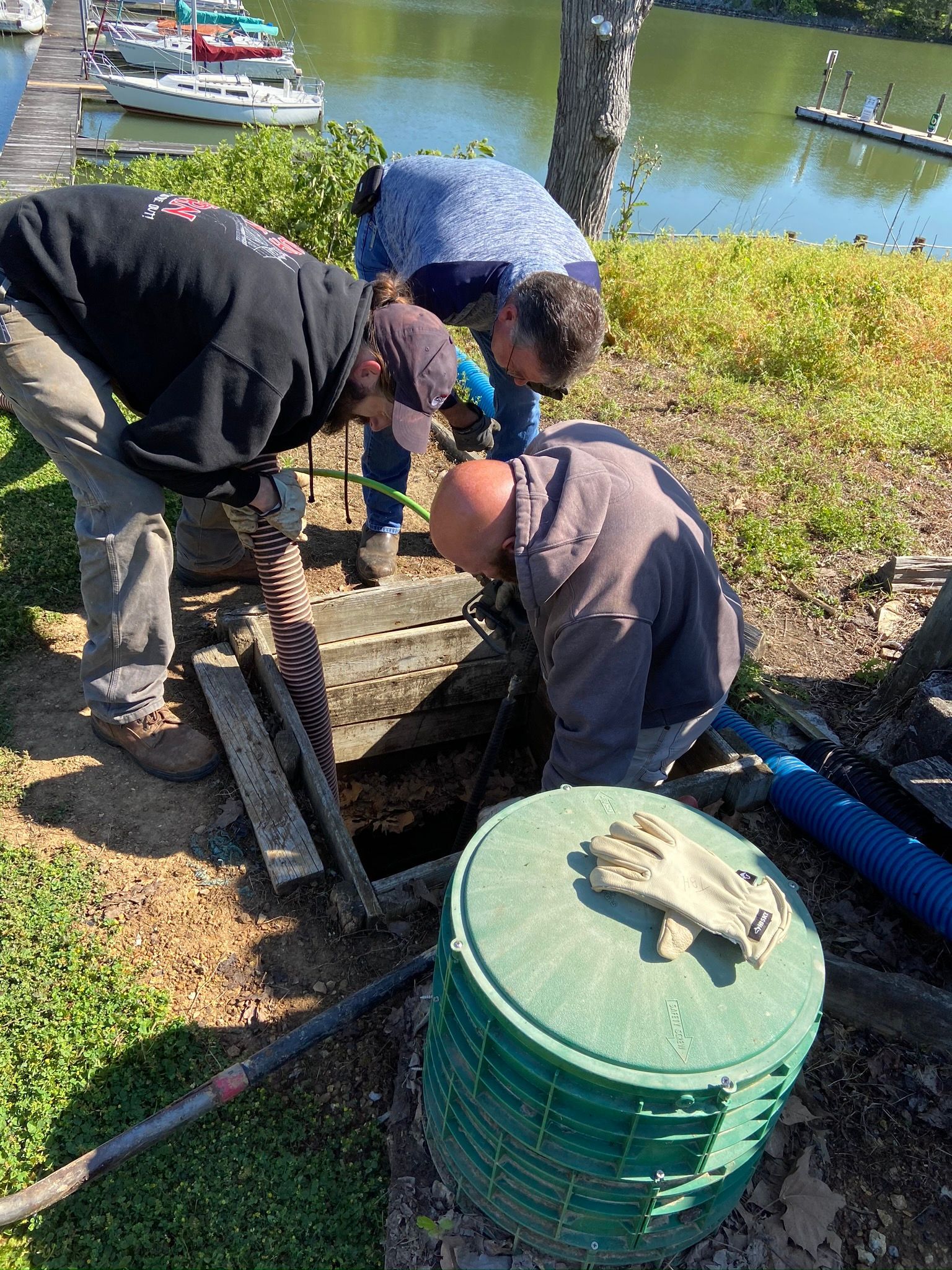 Three people working on an open access point near water and boats. One person kneels, others stand and look in.