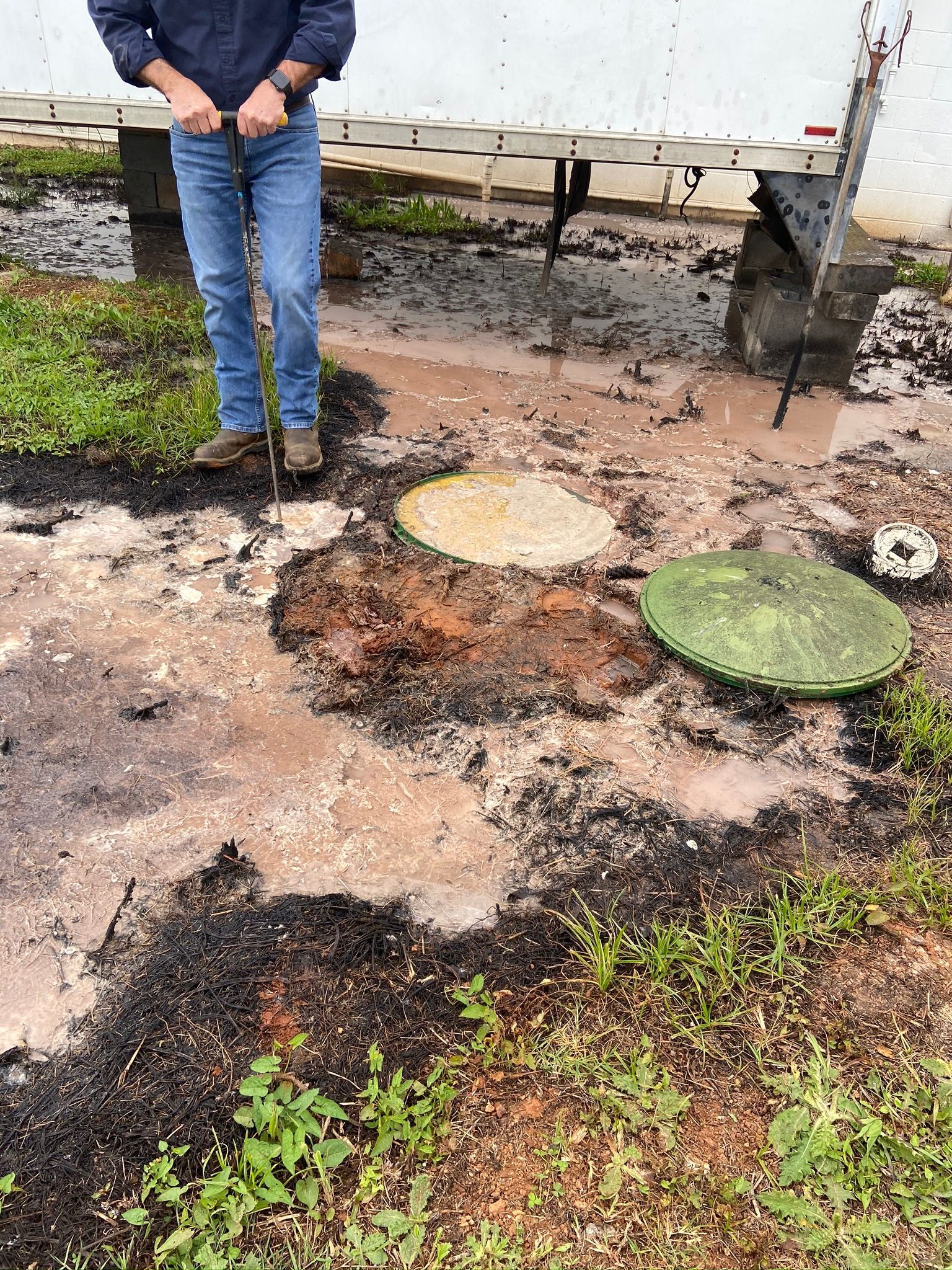Person standing near open septic tank lids in muddy yard.