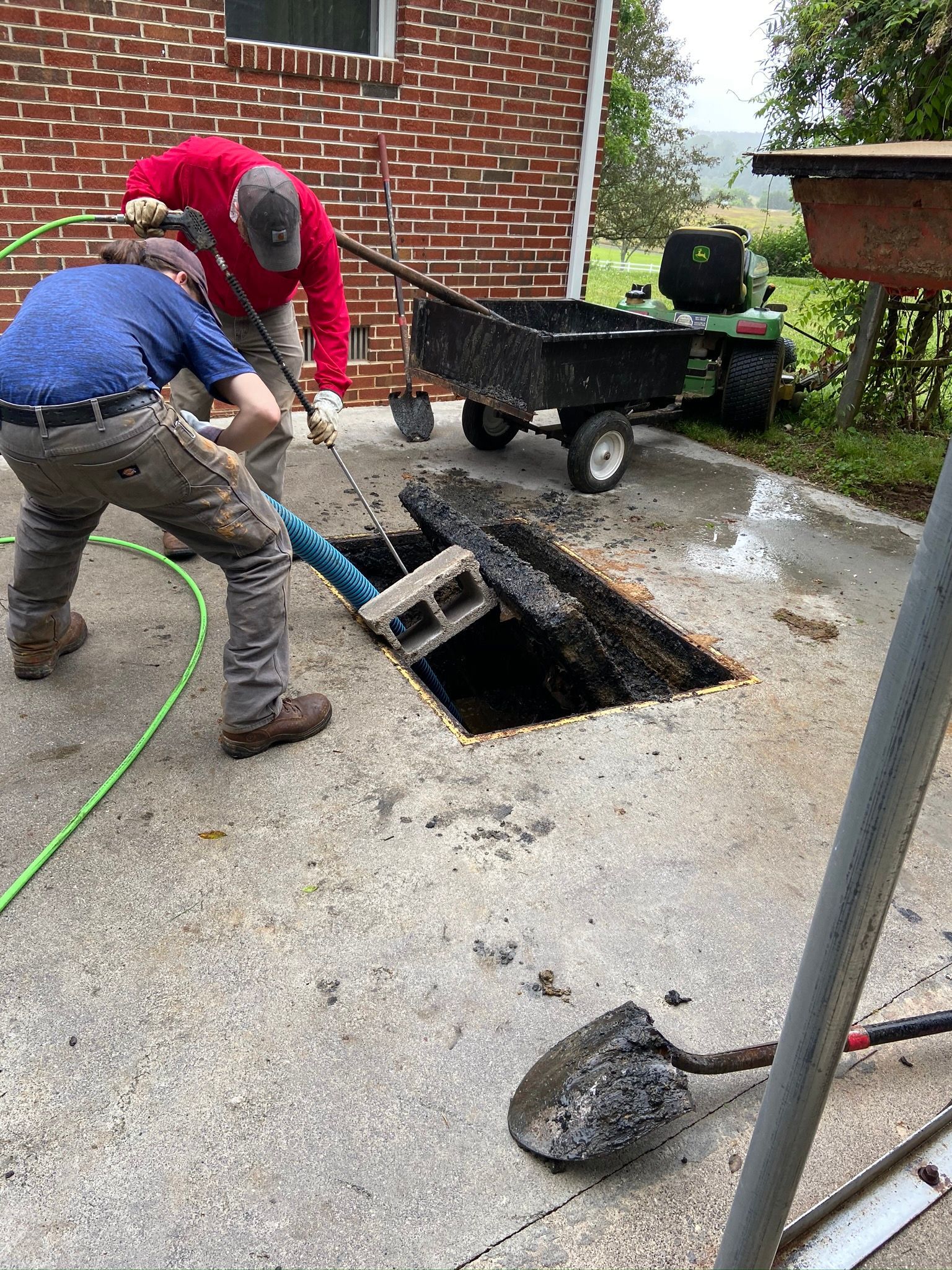 Two people lift a concrete block from a hole in a concrete surface; a wheelbarrow and a John Deere vehicle are nearby.
