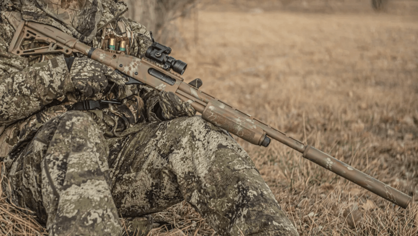 Camouflaged person holding a shotgun outdoors. The gun has a scope and is also camouflaged.