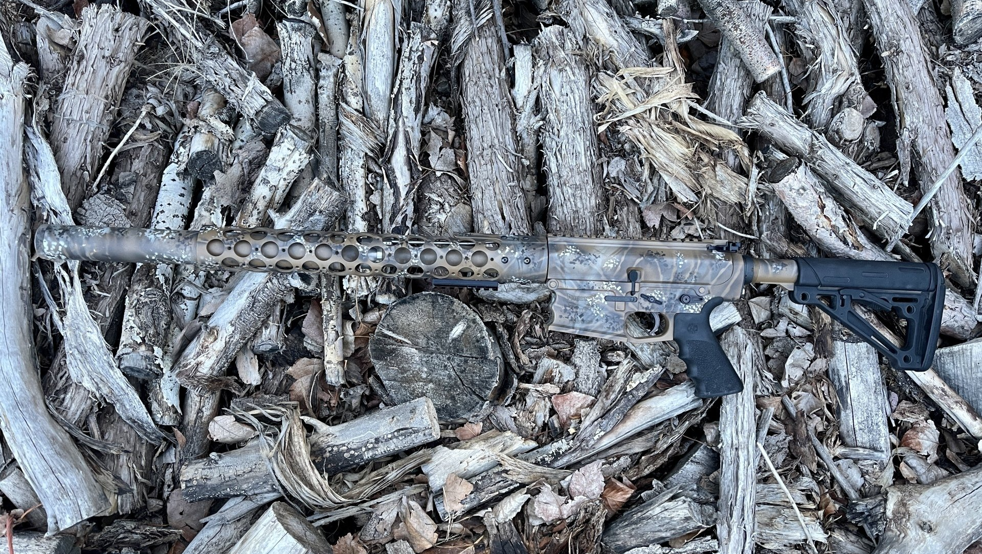 Camouflaged rifle resting on a pile of logs.