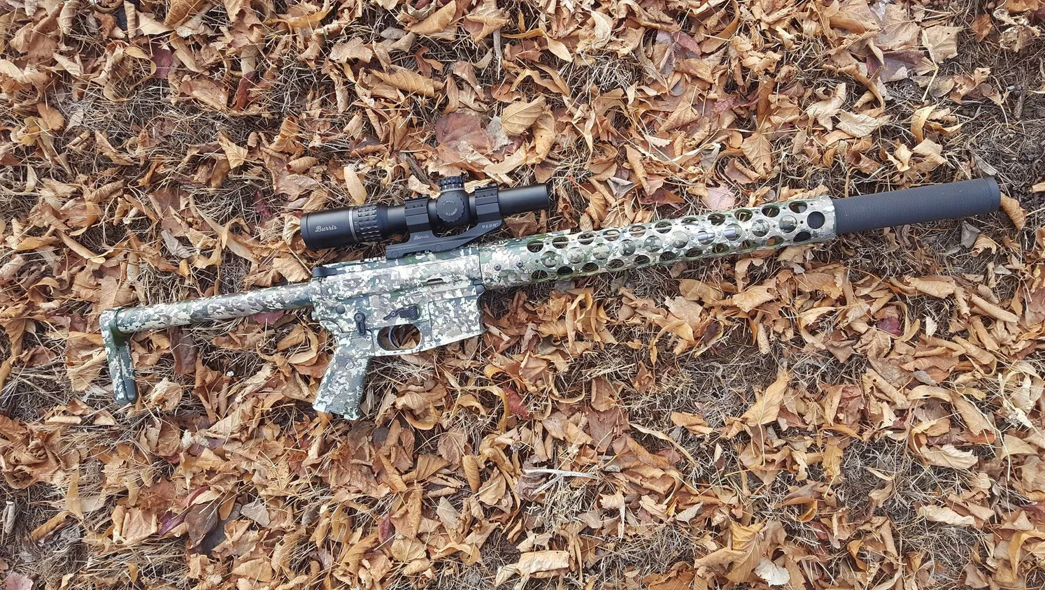 Camouflaged rifle with scope and silencer, lying on brown leaves.