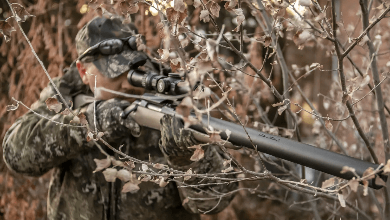 Hunter in camouflage aiming a rifle through foliage.
