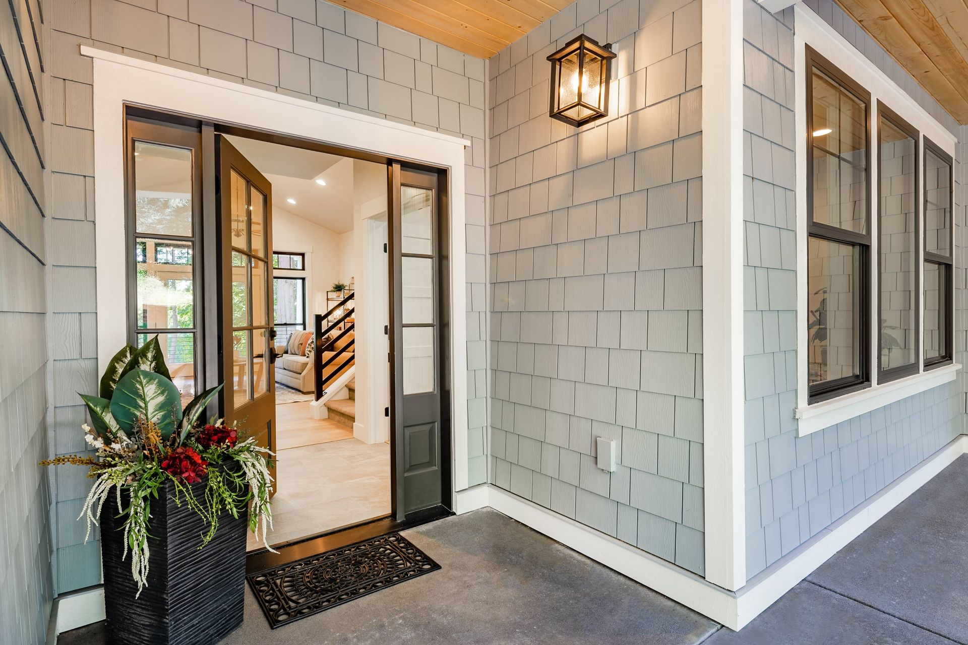Front porch entry with open door, gray shingle siding, lantern light, and potted plants.