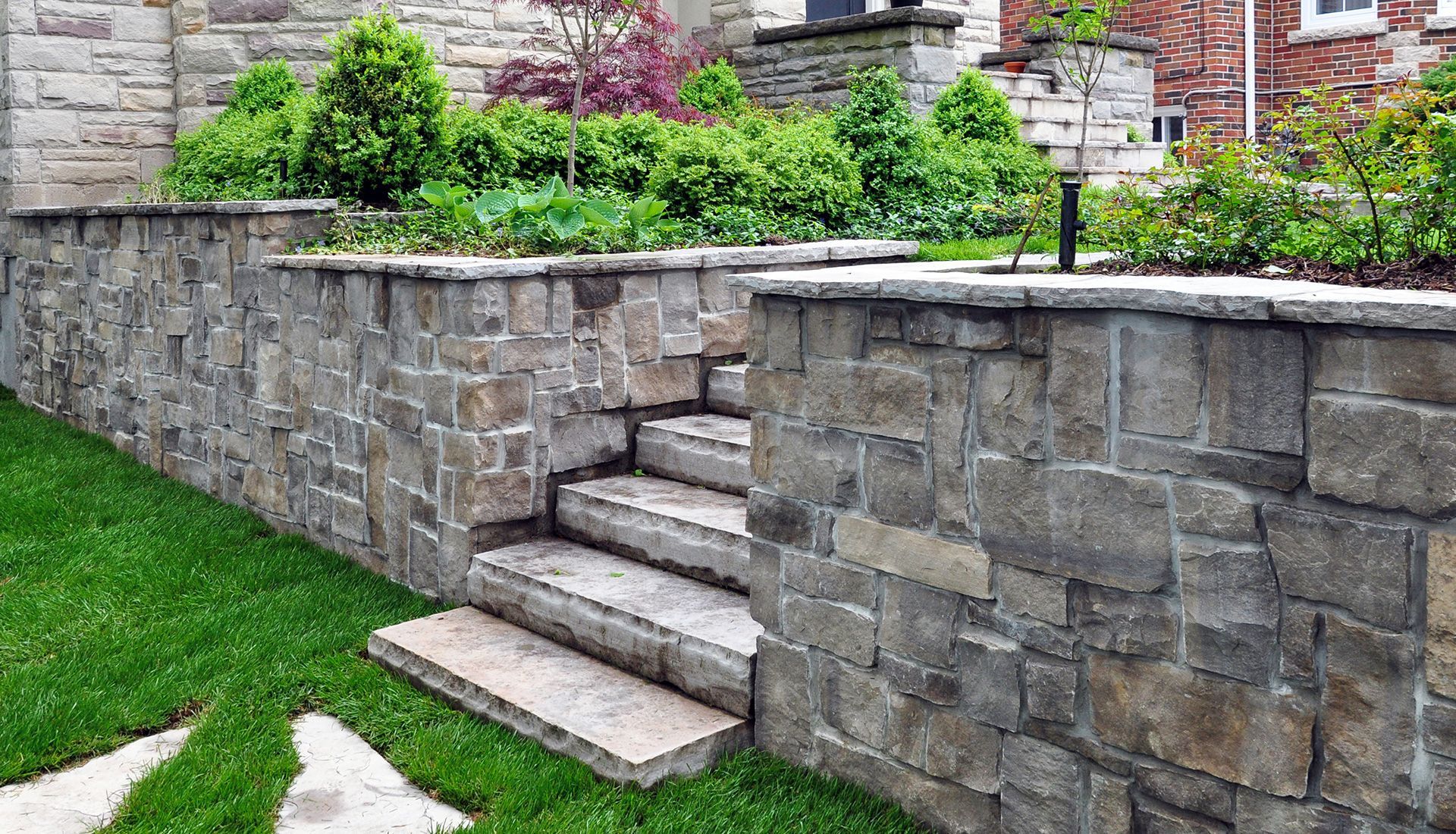 Stone retaining wall with steps leading up to a garden with green shrubs and grass.