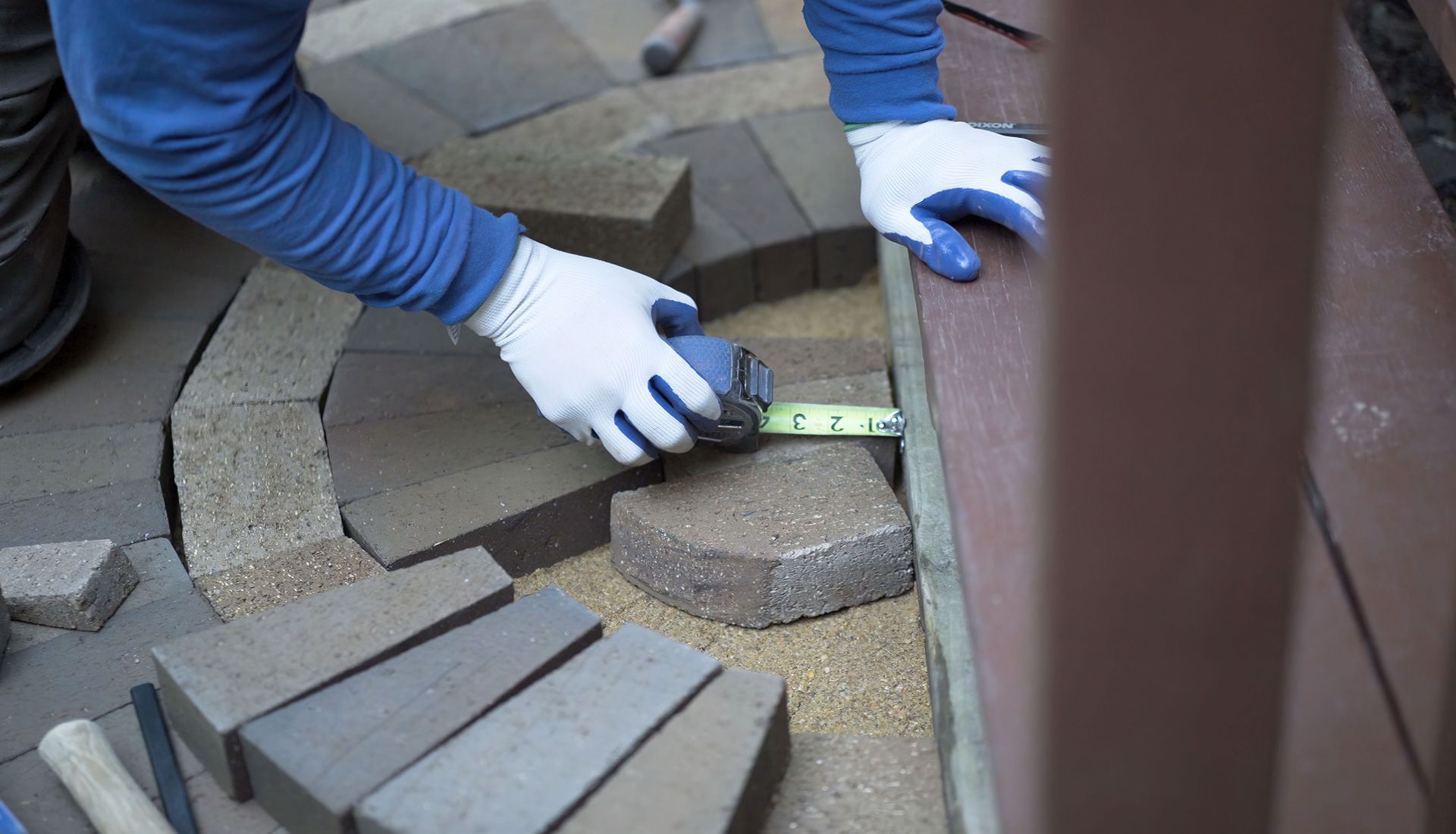 Person in gloves measures paving stones next to a wall, surrounded by other stones and sand.