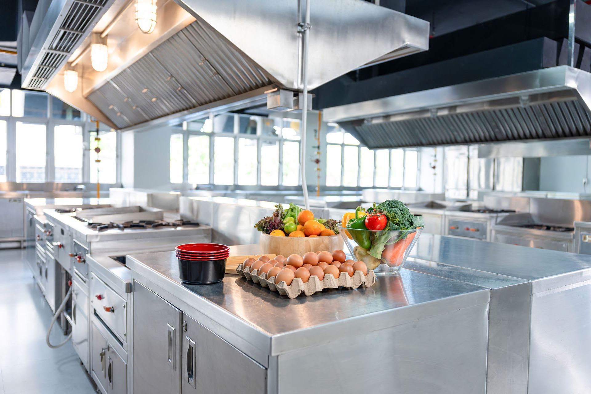 Modern stainless-steel commercial kitchen with prep counters, vents, and food ingredients on the island