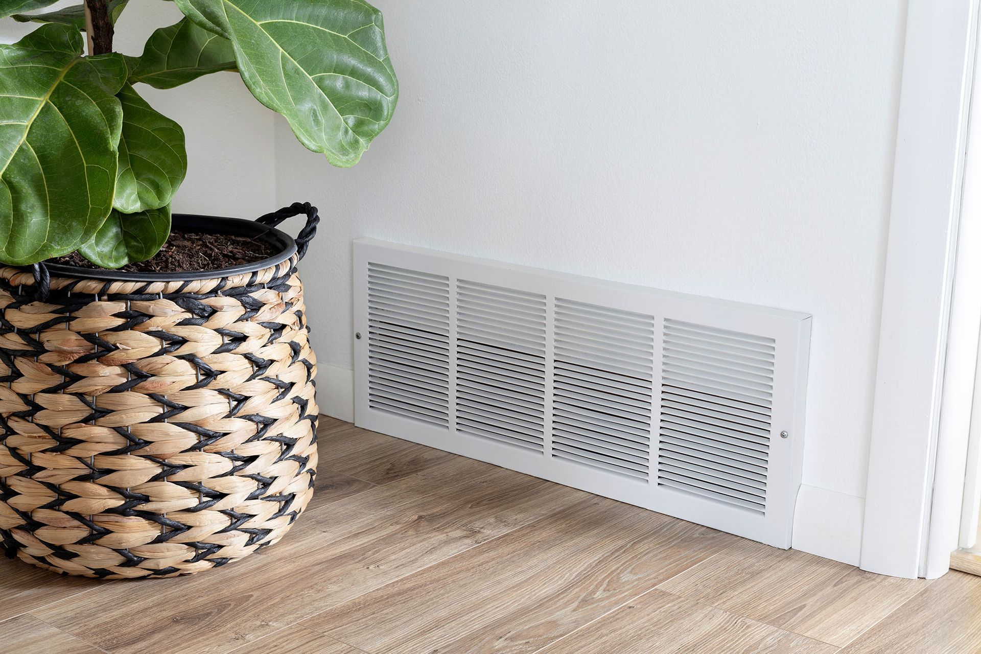 Woven plant basket beside a white wall vent on light wood flooring.