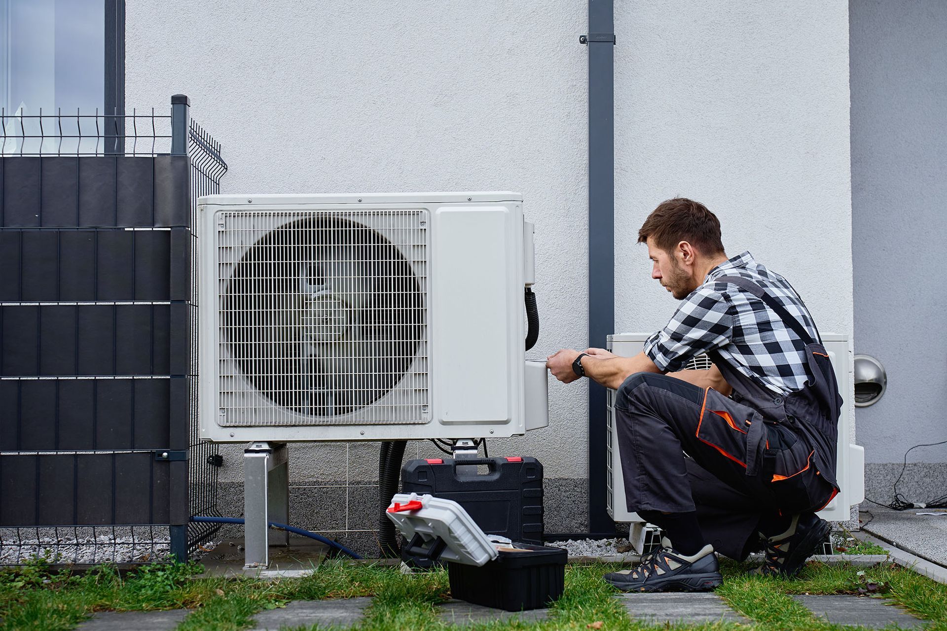 Technician kneels beside an outdoor AC unit, using tools against a gray building wall.