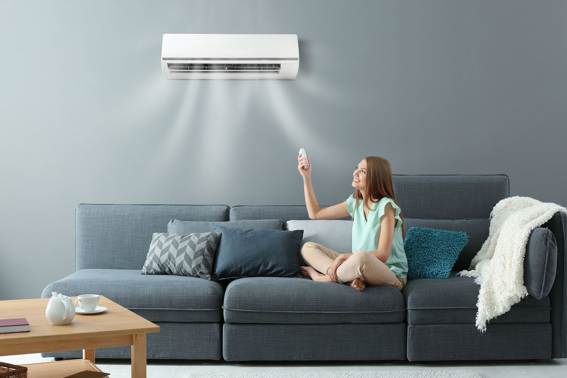 Woman on a gray sofa pointing a remote at a wall-mounted air conditioner in a modern living room
