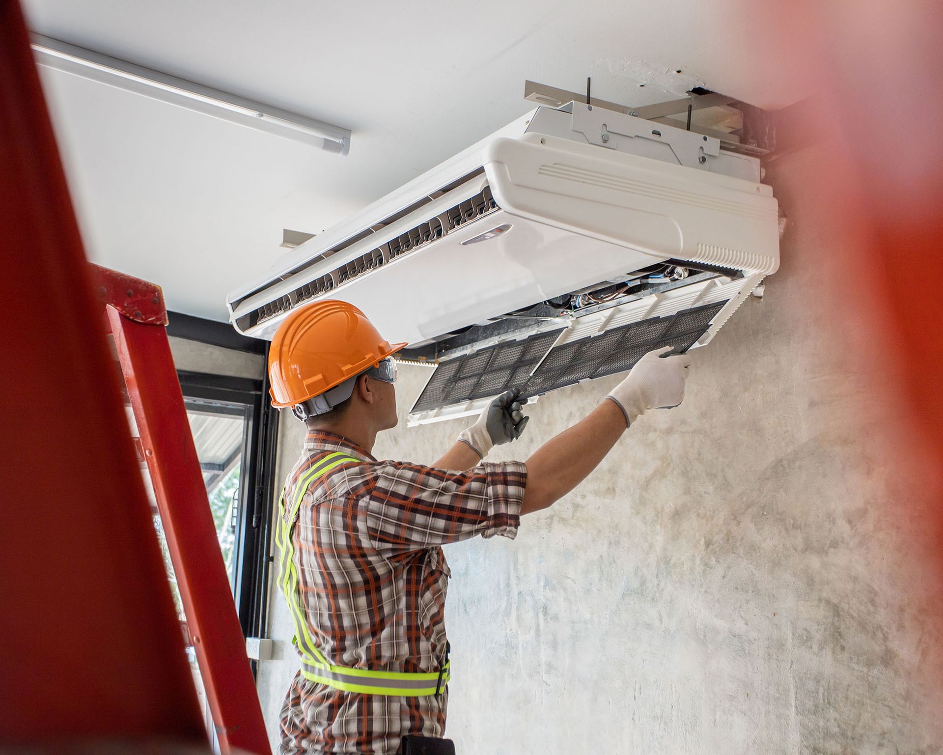 Technician in orange hard hat repairs a ceiling-mounted air conditioner indoors.