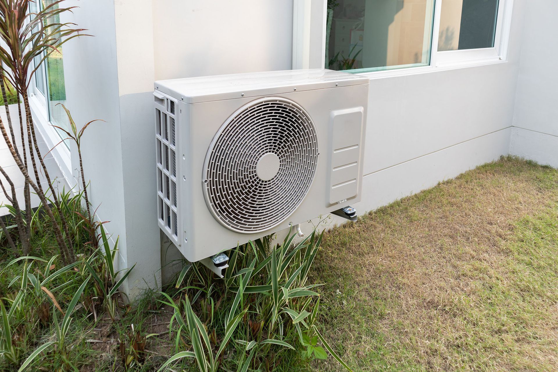 White outdoor air conditioner unit beside a house, next to grass and plants
