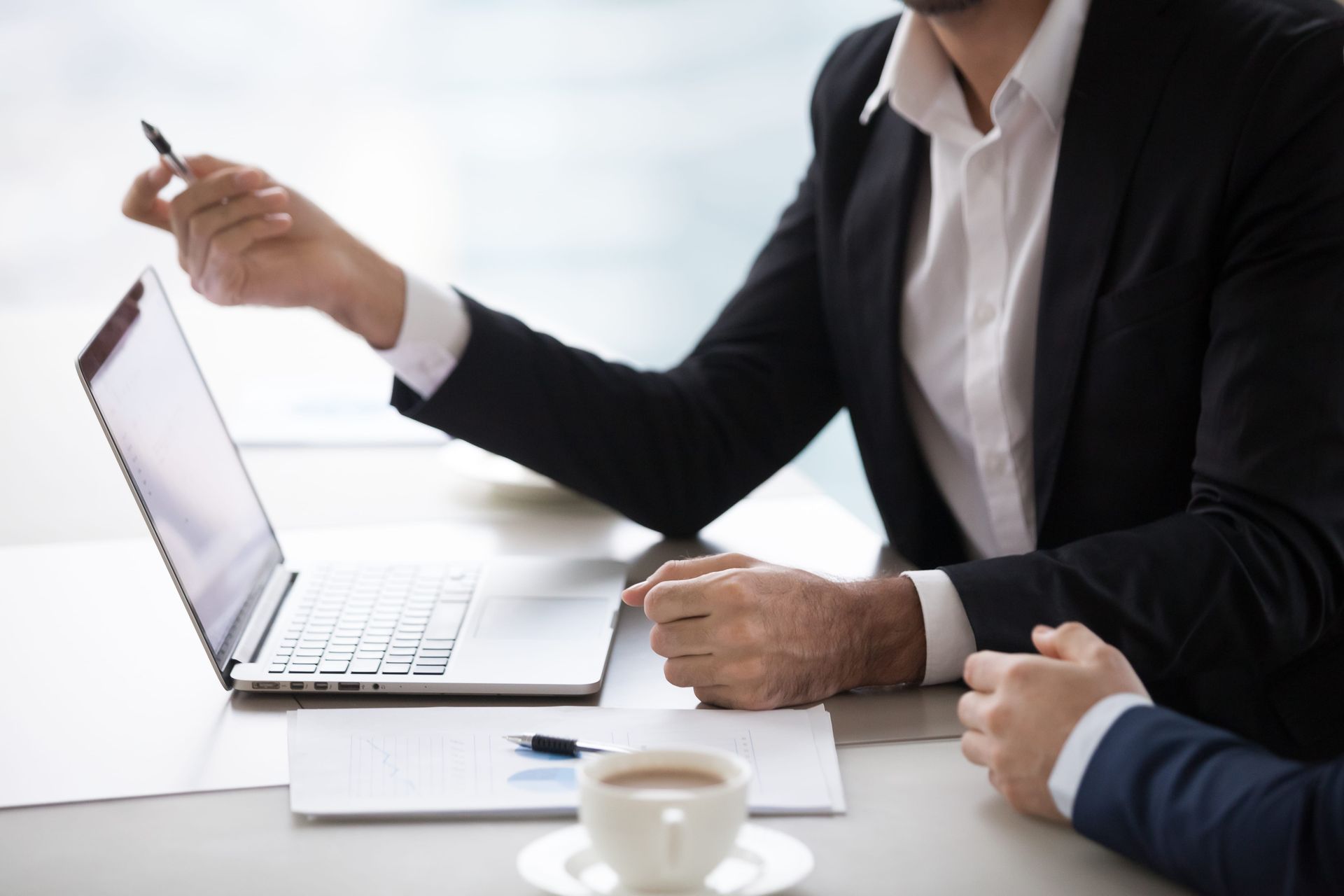 Business meeting at a desk with a laptop, notebook, coffee cup, and two people discussing