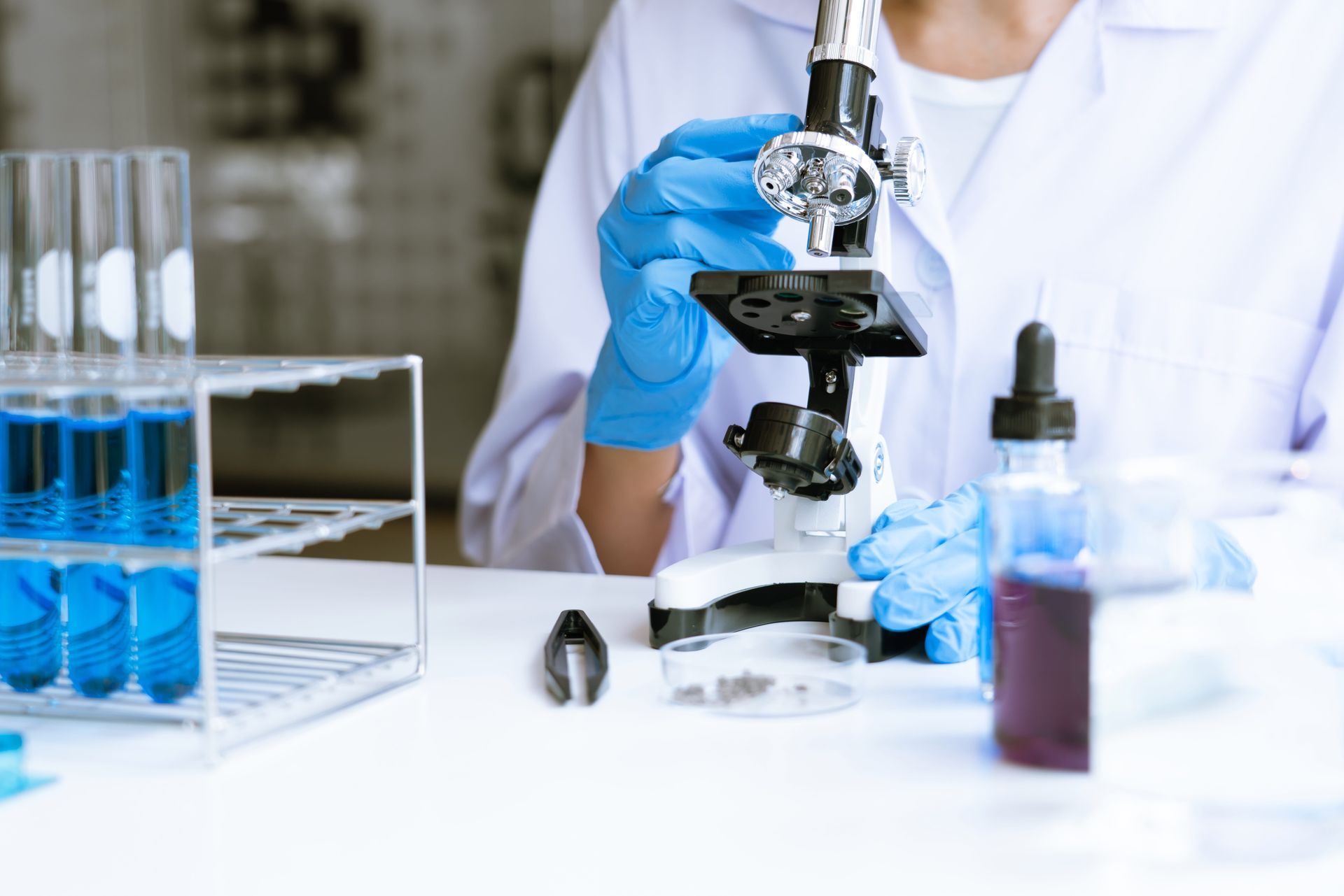 Gloved scientist using a microscope in a lab with test tubes and blue liquid