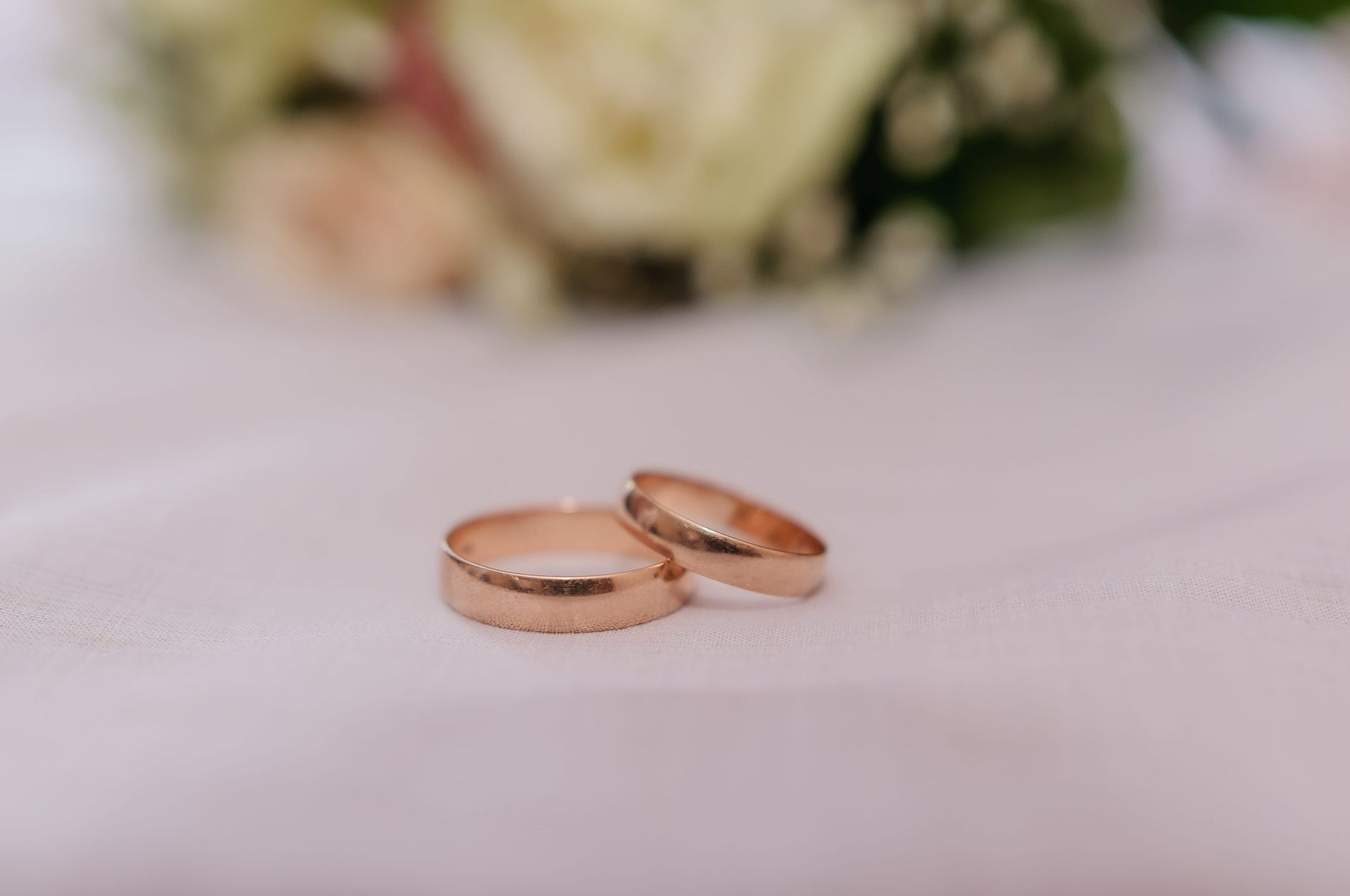 Two gold wedding rings on a soft white surface with a blurred bouquet in the background