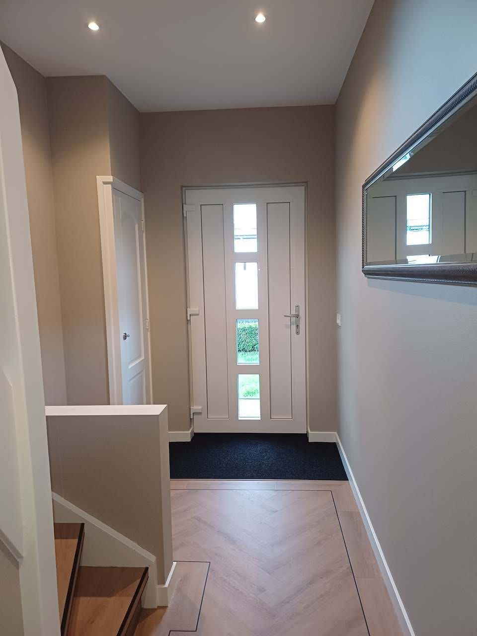 Smalle entreehal met witte voordeur, beige muren, zwarte mat en trapleuning aan de linkerkant.
