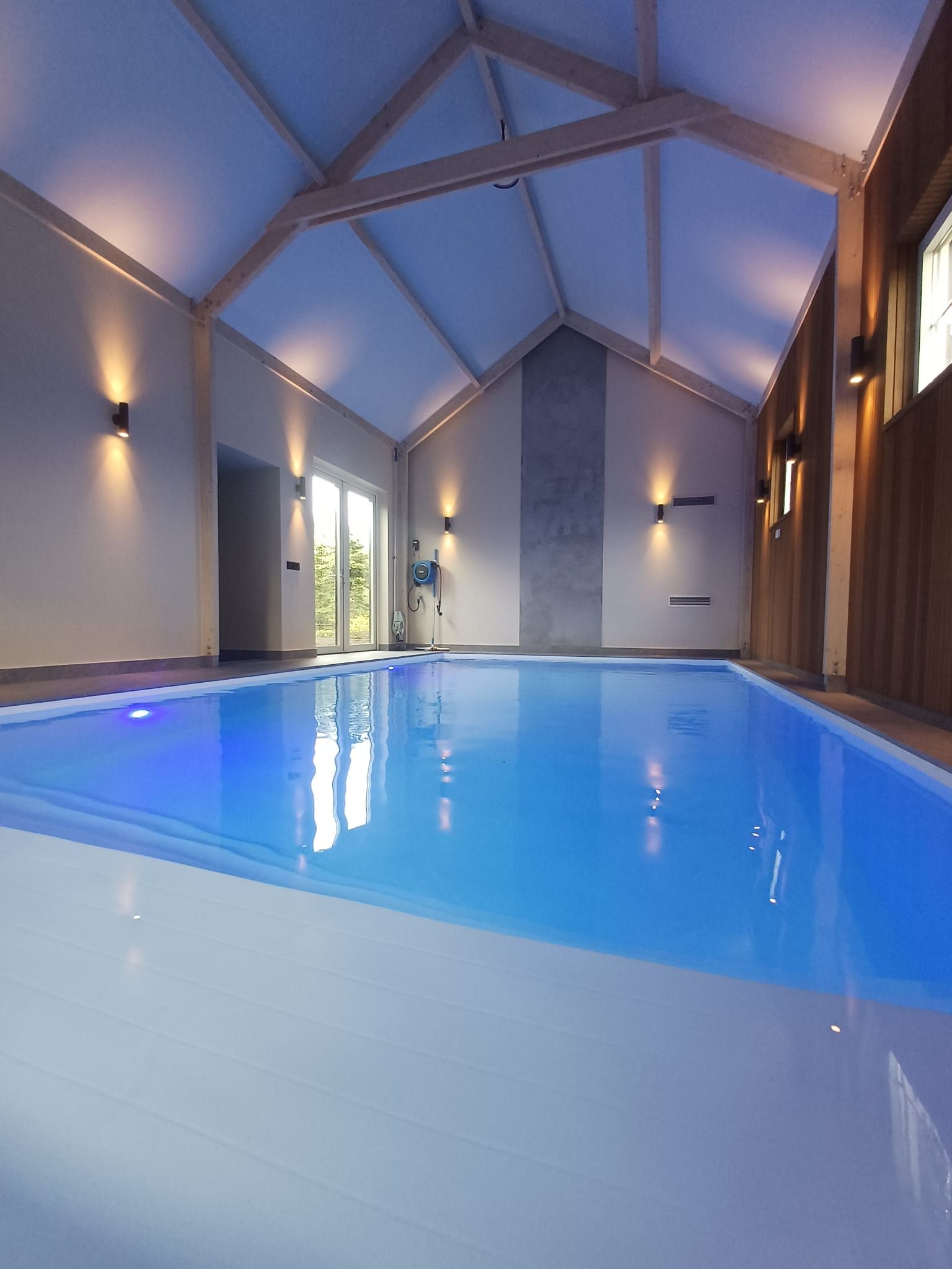 Binnenzwembad met blauw water onder een hoog, geometrisch plafond en warme wandlampen.