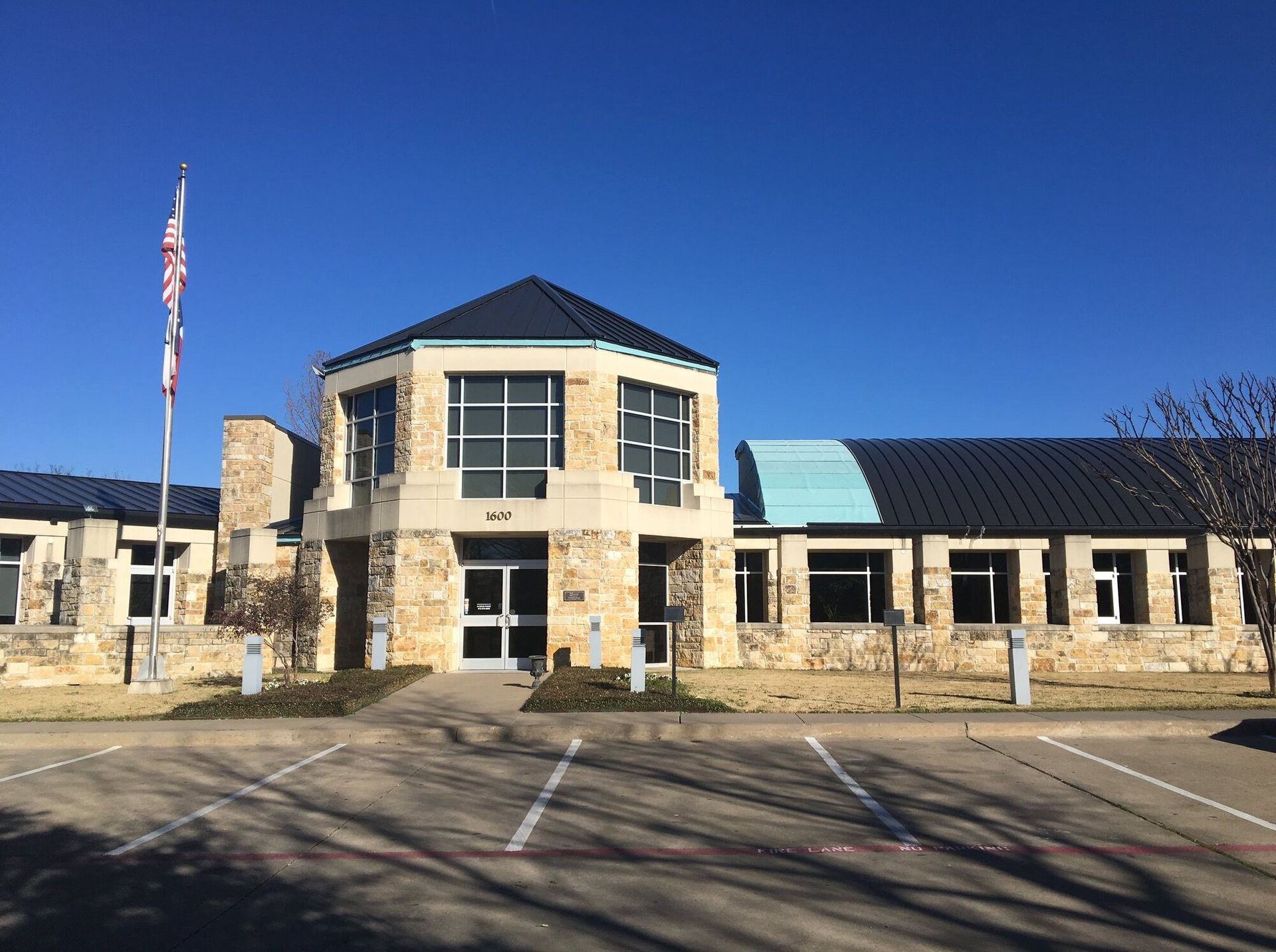 Stone civic building with a central entrance, blue roof accents, and a flagpole under a clear sky