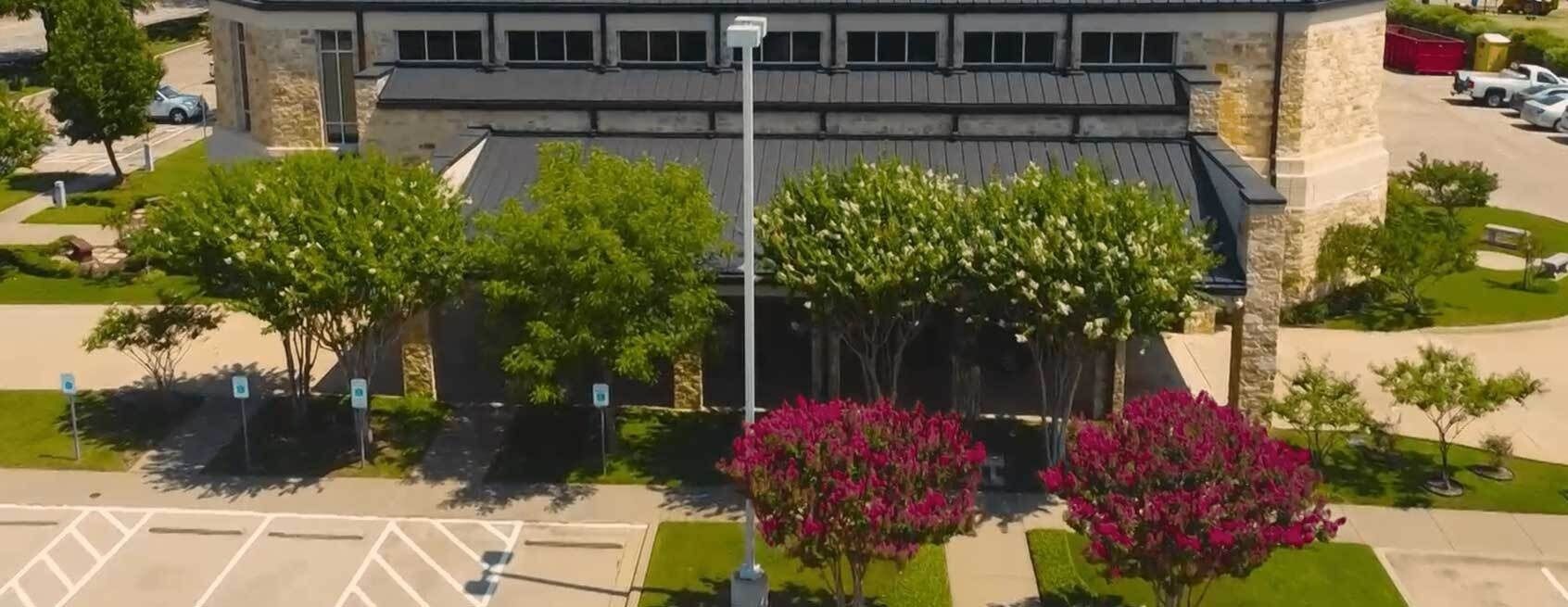 Aerial view of a modern building with tree-lined entrance, parking spaces, and pink flowering shrubs