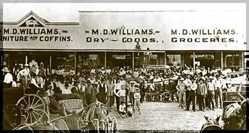 Crowd gathered outside M.D. Williams storefronts with funeral caskets, dry goods, and groceries signs